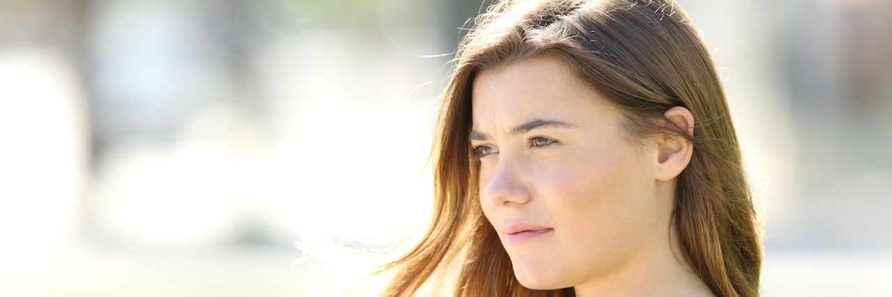 Please Don't Tell Someone With PTSD Their Fear Is 'Illogical' A young woman sitting on a bench outside with a serious expression