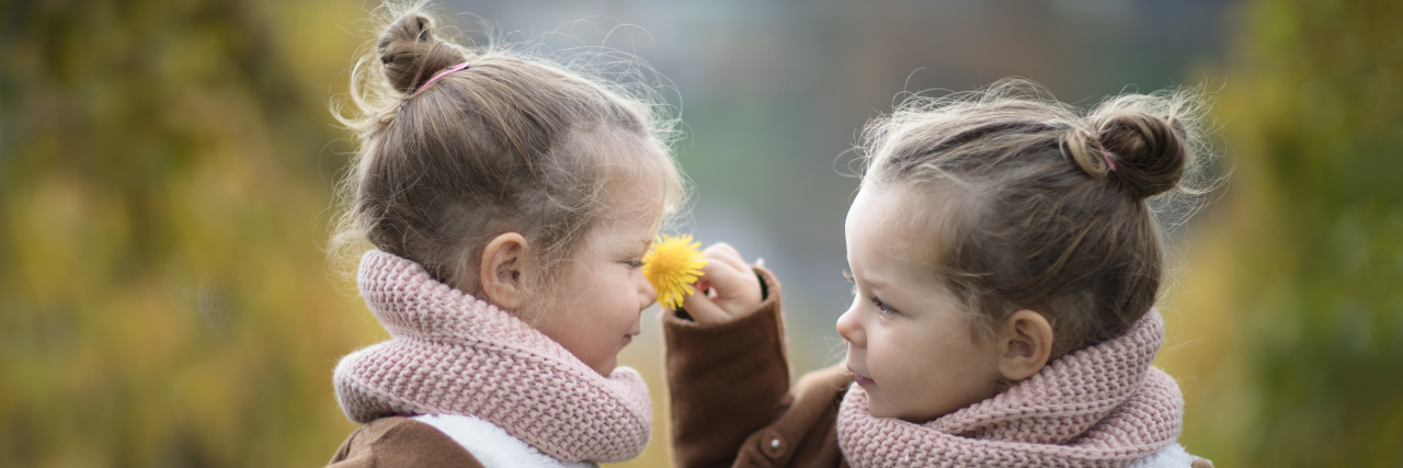 Two little identical twin girls in brown coats smelling a flower.