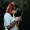 photo of woman with red hair and tattoos checking phone with blurred trees behind her