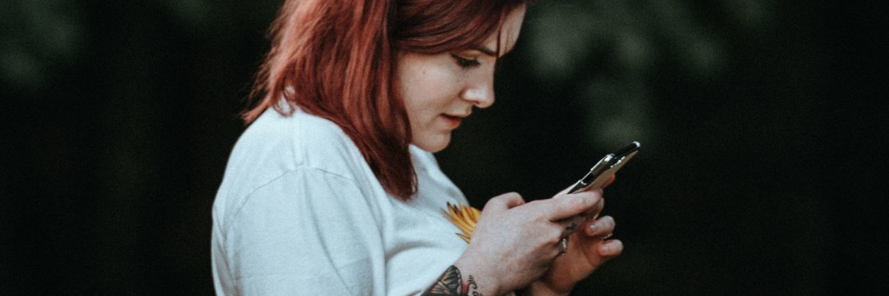 photo of woman with red hair and tattoos checking phone with blurred trees behind her