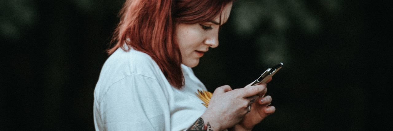 Why Chain Letter Facebook 'Games' About Mental Illness Are Harmful photo of woman with red hair and tattoos checking phone with blurred trees behind her