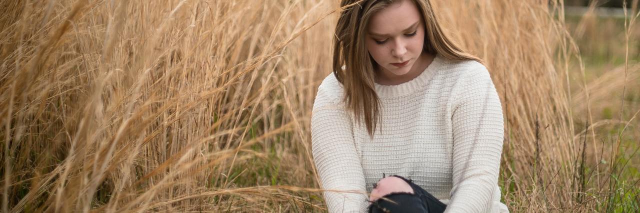 photo of teen girl sitting on ground by long yellowing grass, looking sad