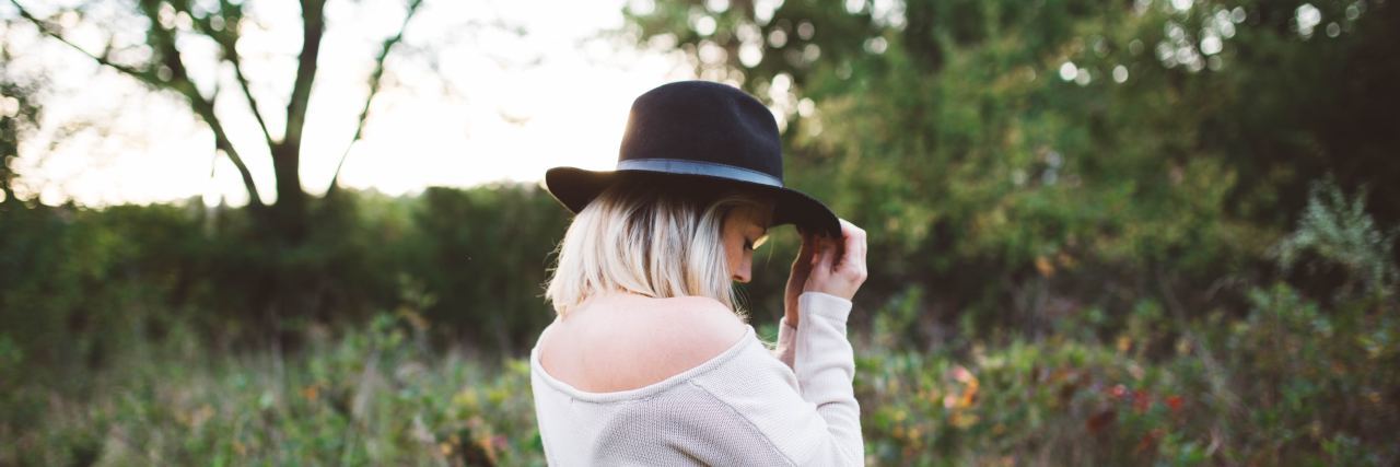 The Media Needs More Stories About Living Successfully With Bipolar woman outside in a field wearing a off-white sweater and black hat over her blonde hair, her hand holding the top of the hat