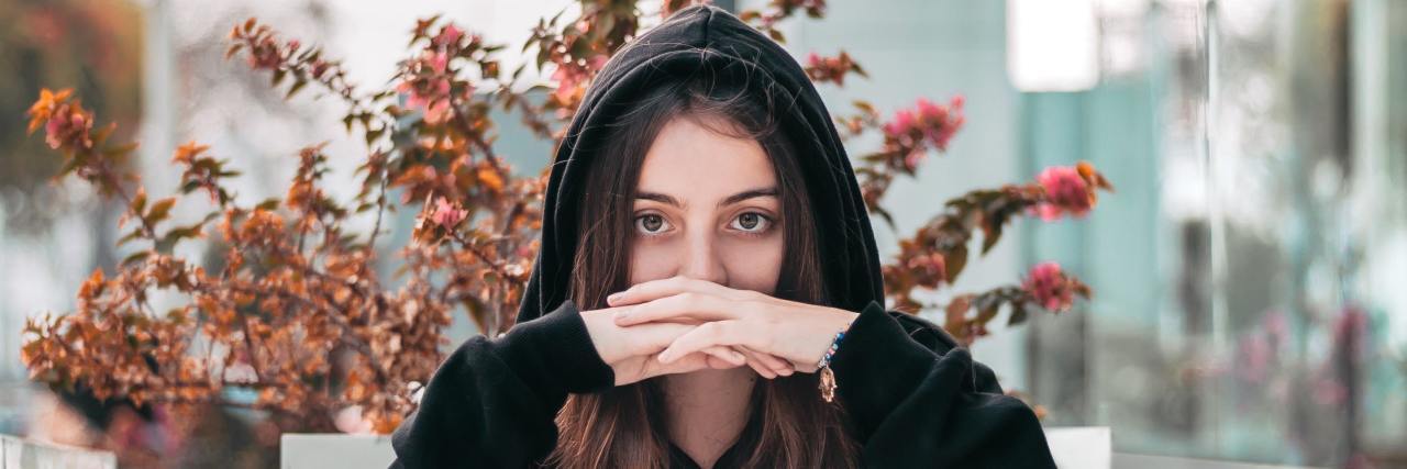 Stop Joking About OCD and Using It as an Adjective woman in a black hoodie with her hands folded, covering her mouth staring straight into the camera seriously