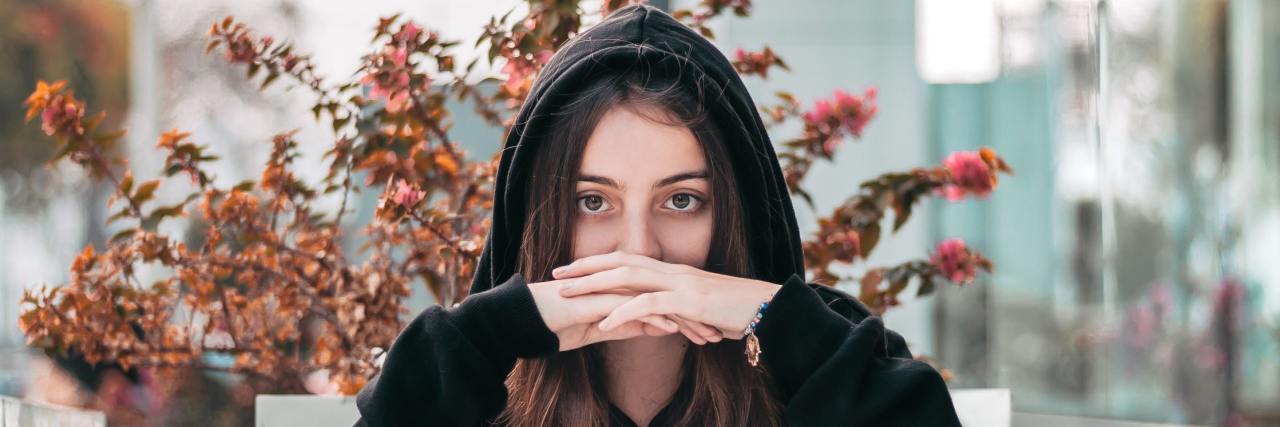 woman in a black hoodie with her hands folded, covering her mouth staring straight into the camera seriously