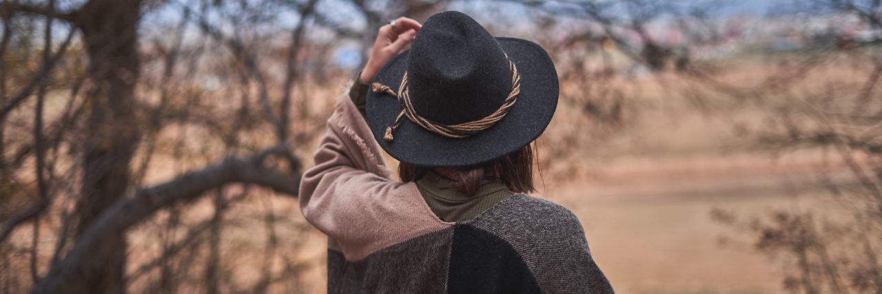 Recovery From Bipolar Disorder Takes a Combination of Things woman facing back of camera wearing a black hat and cardigan, arm raised, walking outside in a dessert