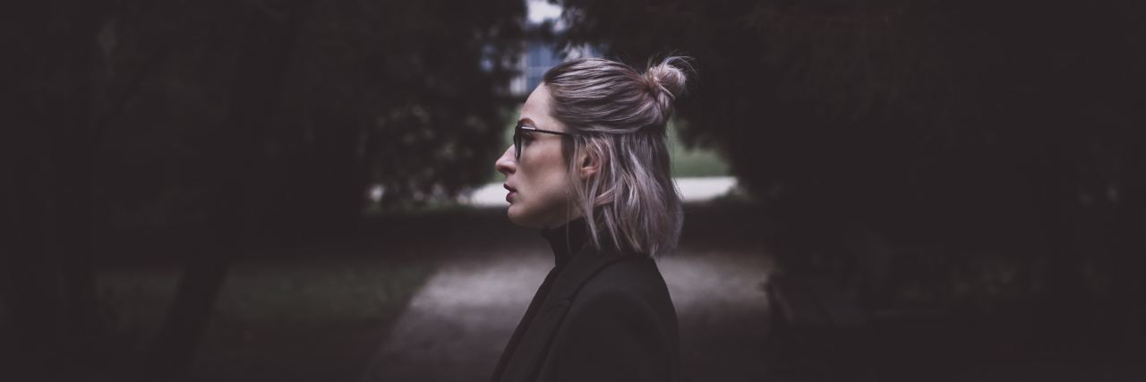 woman standing on a path in a forest looking sideways with glasses on and her hair in a bun with a straight face