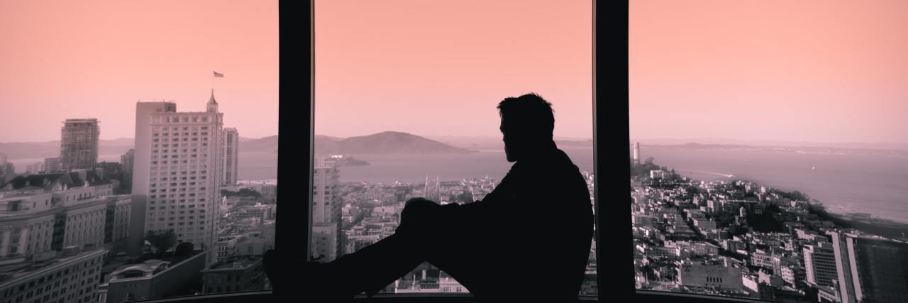 man sitting on a bench in a building looking outside the window at the city during sunset