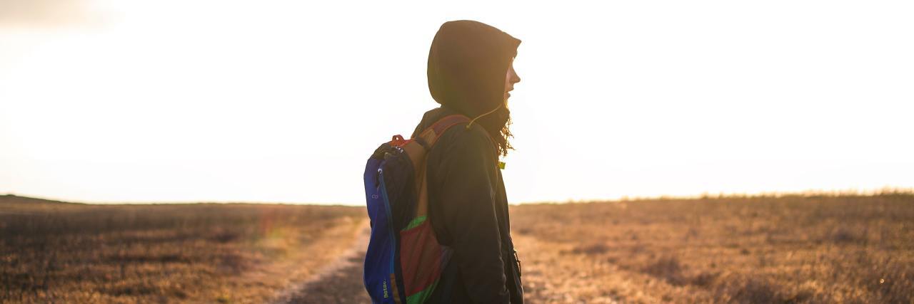 person standing on a gravel road looking to the side wearing a backpack and a black jacket with the hood up