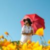 photo of blonde woman in white dress standing in field of yellow flowers, holding a red umbrella under blue skies
