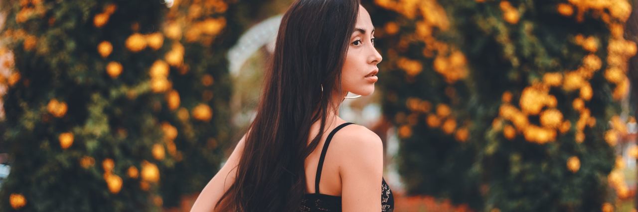 We Need to Stop Looking for ‘Healing’ Like It’s a Destination photo of woman walking down tree and flower lined road looking over shoulder