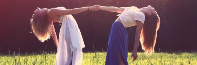 photo of two women in meadow holding each other's hands, facing each other and leaning back away from each other