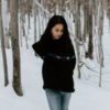 photo of young woman standing in snowy woods alone and looking sad, arms wrapped around herself