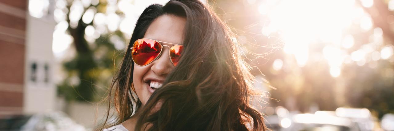 Woman with brown hair wearing sunglasses, smiling with the sun shining through the trees behind her.