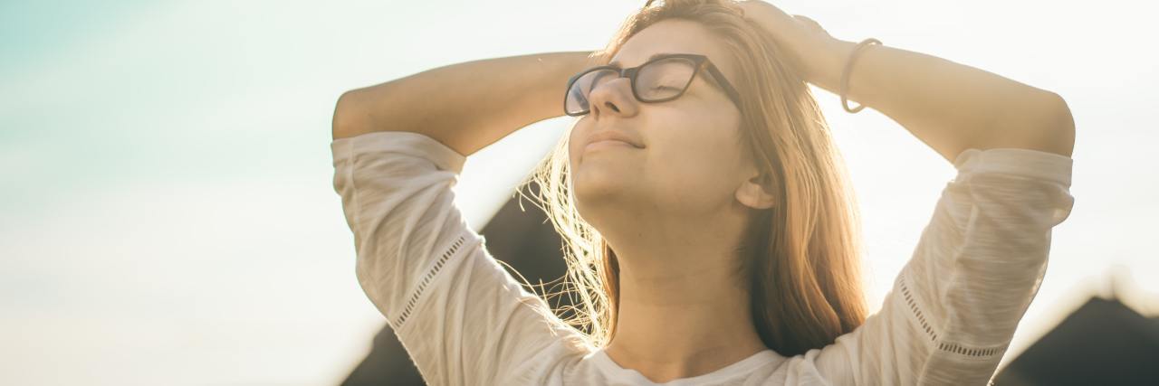 Why Self-Care Isn't Selfish young woman in glasses standing outside with her head back, eyes closed, embracing the sun
