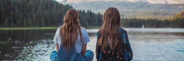 2 girls with their backs to the camera sitting on a rock ledge overlooking a lake with a mountain in the distance