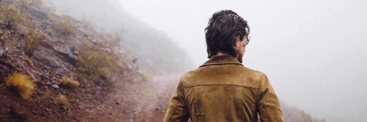 back of a slim man on a dirt path with his hands in a leather jacket
