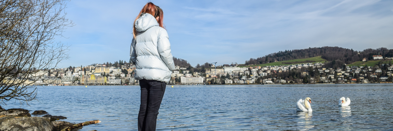 How I Travel Despite Having Crohn's Disease photo of contributor - young woman standing on shore with an exotic town in the distance and swans in front of her
