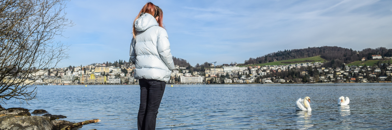 photo of contributor - young woman standing on shore with an exotic town in the distance and swans in front of her