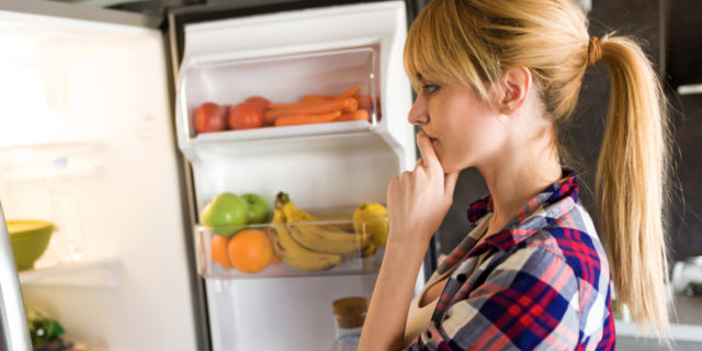 A young woman is looking in the fridge hesitantly.