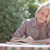 older woman in flowered shirt sitting outside writing