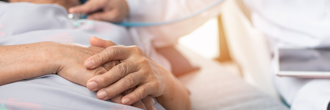 COVID-19: How Do You Treat the Coronavirus? Hospitalized elderly patient, senior old aging woman laying on bed with cardiologist doctor or physician examining cardiological heart health, checking pulse in hospital clinic exam room