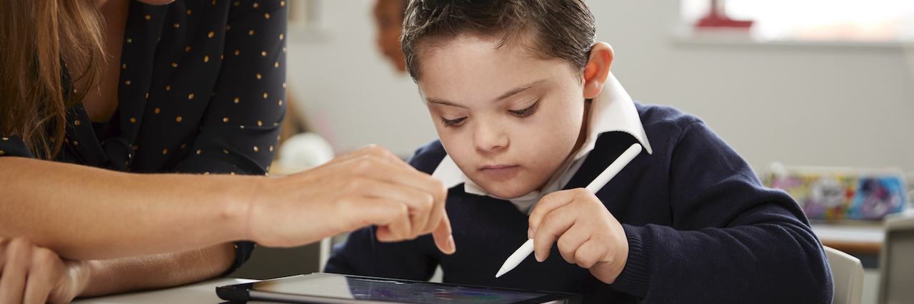 Senate's COVID-19 Bill Proposes Review of School for Disabled Students Young female teacher working with a Down syndrome schoolboy sitting at desk using a tablet computer in a primary school classroom, front view, close up