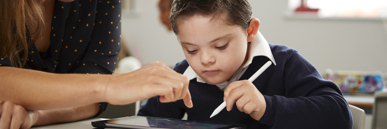 Young female teacher working with a Down syndrome schoolboy sitting at desk using a tablet computer in a primary school classroom, front view, close up