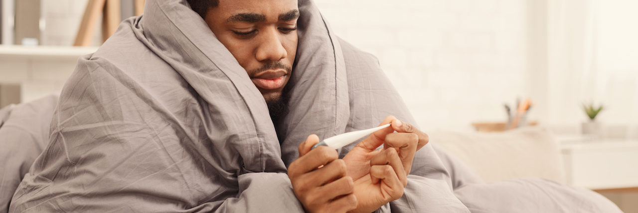 What's the Difference Between COVID-19 and the Flu? Ill african-american man covered with blanket holding thermometer, having fever, copy space