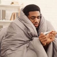 Ill african-american man covered with blanket holding thermometer, having fever, copy space