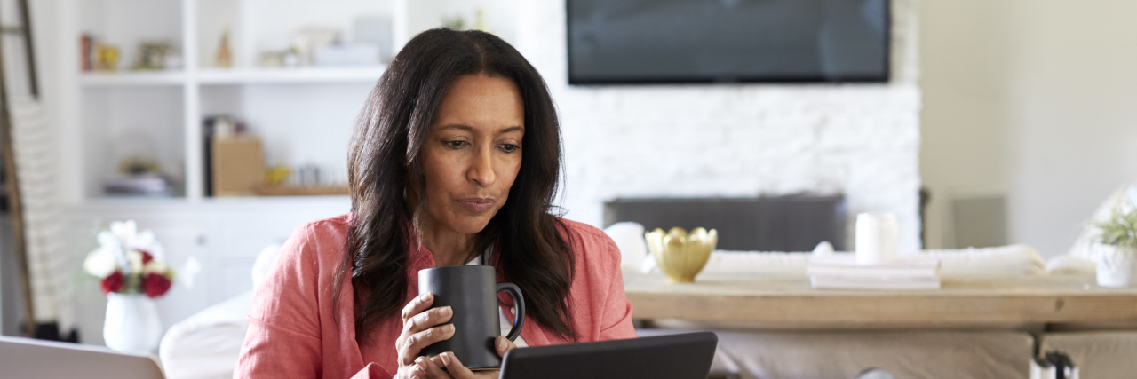 Why Working Remotely Can Be Challenging When You Have De Middle aged woman sitting at a table reading using a tablet computer, holding a cup, front view