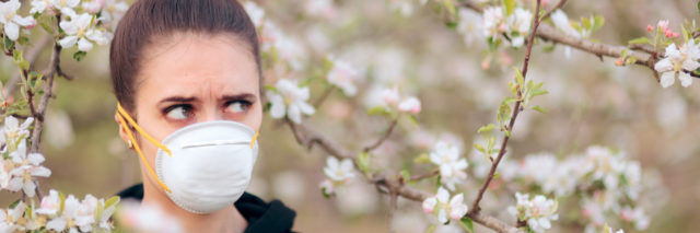White woman with brown hair in a bun wearing a face mask and standing among white flowering trees