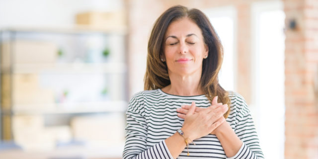 woman in a striped shirt standing in a room with her eyes closed and hands over her heart