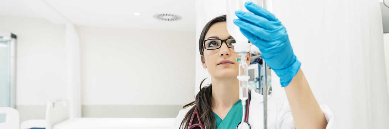 COVID-19: Pharmacy Staff Who Prepare IV Drugs Face Mask Shortages Young woman doctor anesthesiologist dressed in green gown, prepares an IV in hospital room