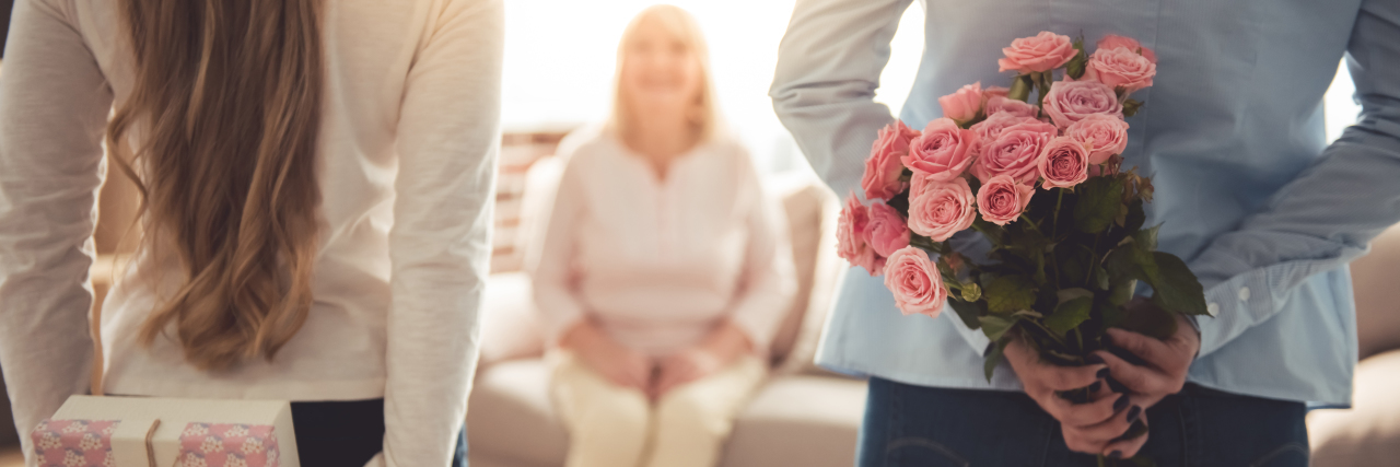 mother and daughter with presents behind their backs, getting ready to surprise the grandmother sitting on the couch