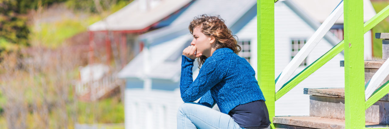 Why Going Home Is Hard for Some Students During Coronavirus Outbreak Young woman sitting on porch of colorful painted house in idyllic countryside summer scenery in Scandinavia or Canada with hand on face thinking