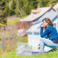 Young woman sitting on porch of colorful painted house in idyllic countryside summer scenery in Scandinavia or Canada with hand on face thinking
