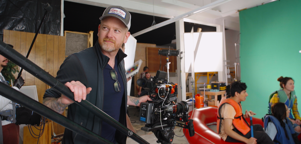 'Making Sense' director Greg Bayne, a white man with a redish beard wearing a ball cap and dark blue shirt, standing behind a camera on set