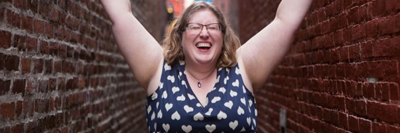 photo of Lindley Ashline, body positive icon and owner of Body Liberation Stock and The Body Love Box. She is seen in an alley, smiling with her eyes closed and raising her arms high