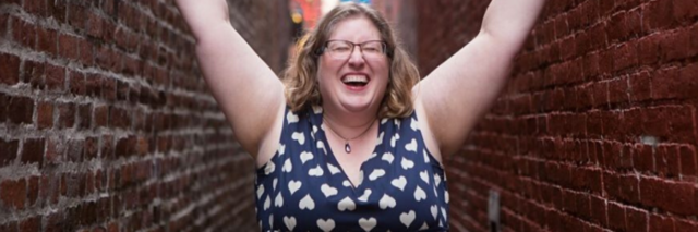 photo of Lindley Ashline, body positive icon and owner of Body Liberation Stock and The Body Love Box. She is seen in an alley, smiling with her eyes closed and raising her arms high
