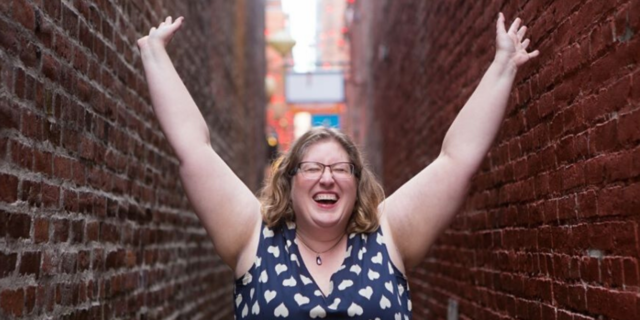 Interview: What Body Positivity Icon Lindley Ashline Wants You to Know About Your Self-Image photo of Lindley Ashline, body positive icon and owner of Body Liberation Stock and The Body Love Box. She is seen in an alley, smiling with her eyes closed and raising her arms high