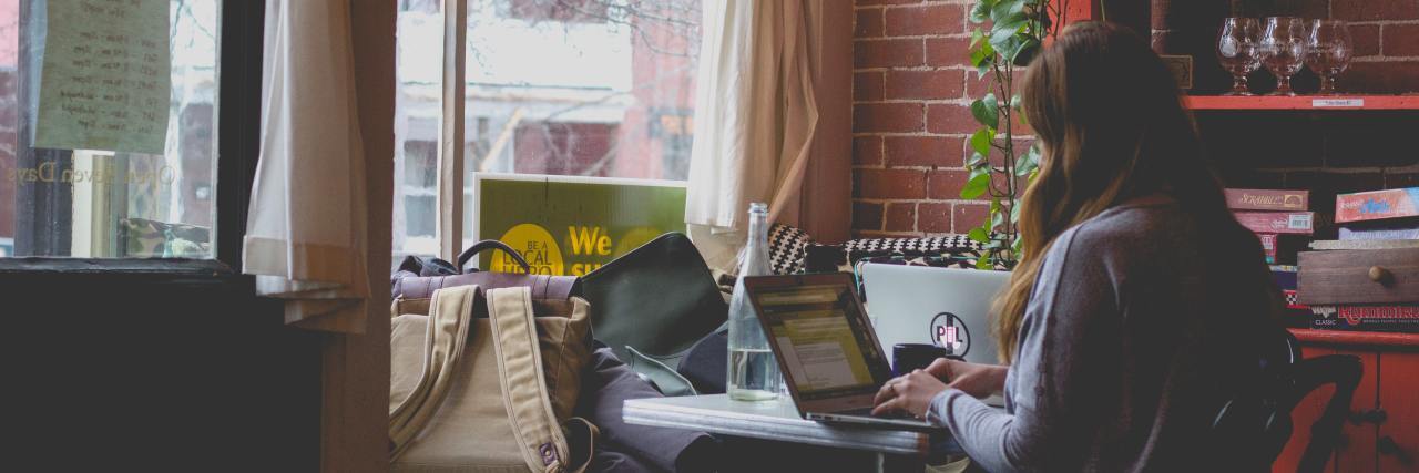 How Job Hunting With Anxiety Leads Me to Fantasize About a New Life woman in a cozy apartment sitting at a table working on her computer