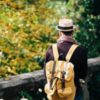 photo of man in hat and yellow backpack standing on bridge with trees
