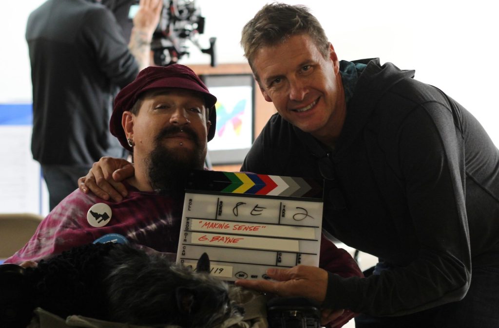 Doug Cole (a white man wearing a black shirt) holding a movie slate next to quadriplegic Ahniah Selene, a white main with a long, brown beard and a maroon hat and shirt