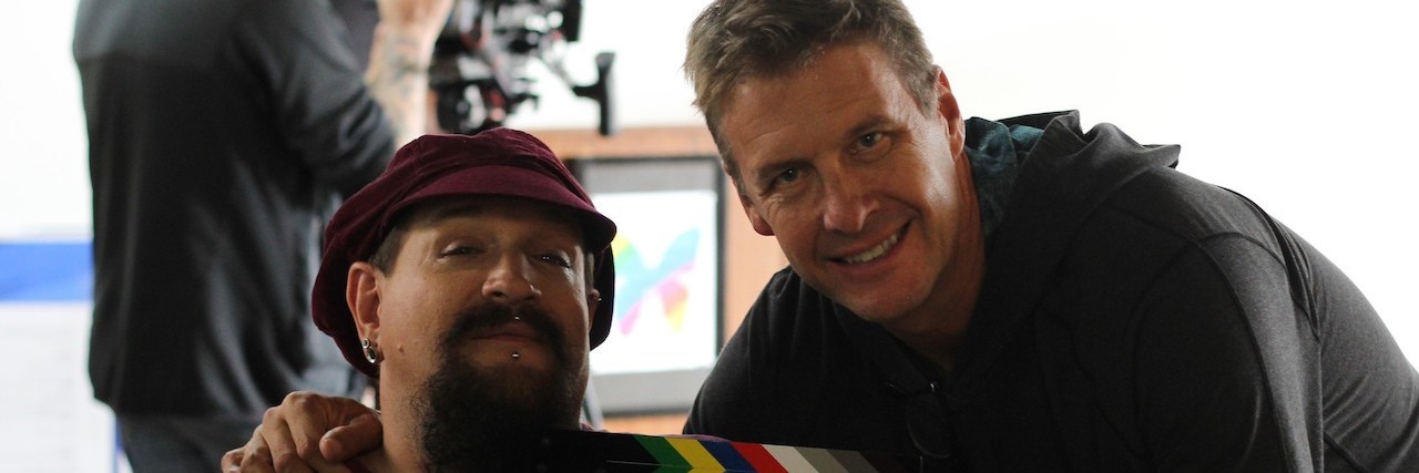 'Making' Sense' Sci-Fi Action Film Features Actors With Disabilities Doug Cole (a white man wearing a black shirt) holding a movie slate next to quadriplegic Ahniah Selene, a white main with a long, brown beard and a maroon hat and shirt