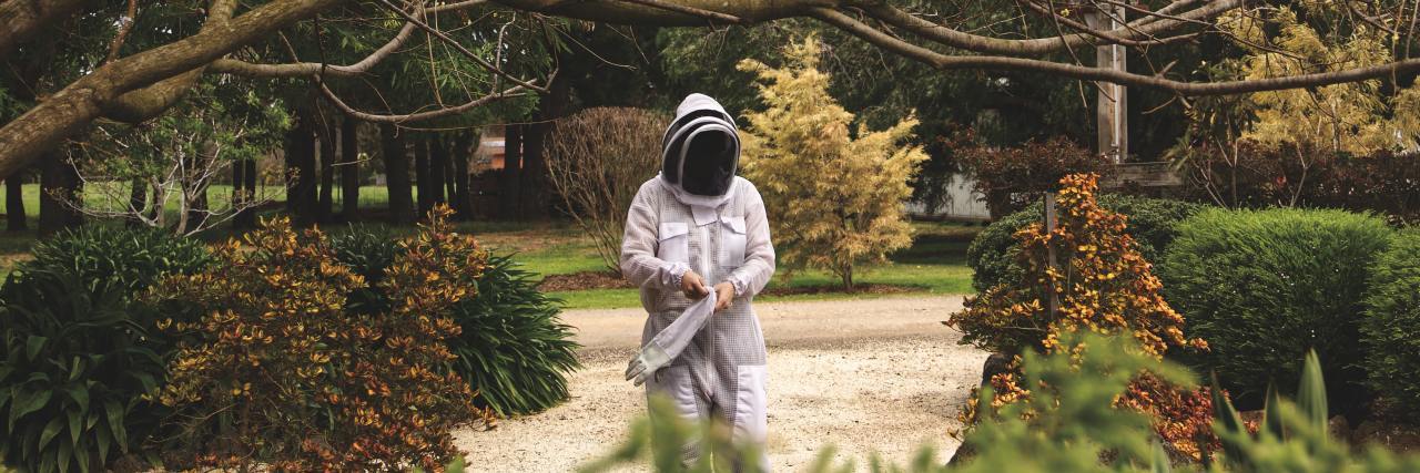 photo of person in white beekeeping outfit in middle of path with trees