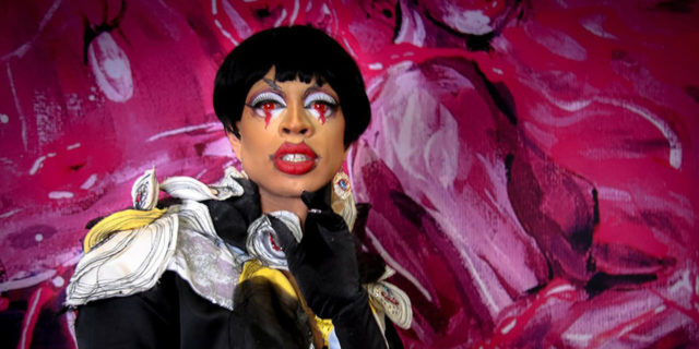 Yvie Oddly poses while in drag in front of a pink wall