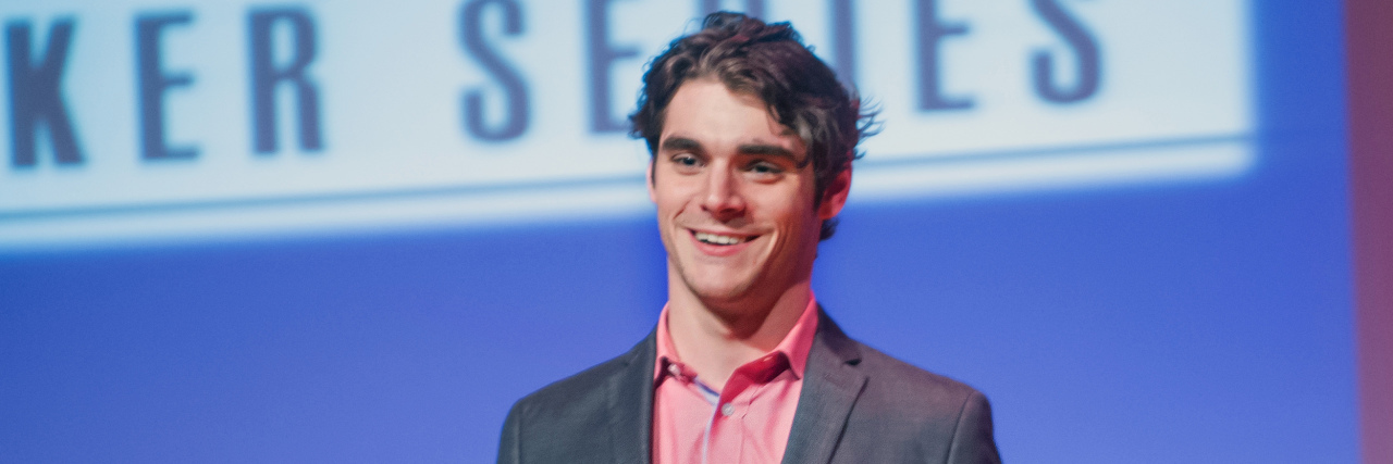 RJ Mitte speaks onstage in a grey suit at an event