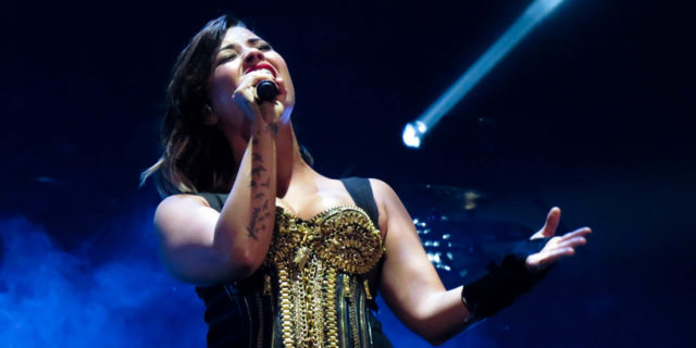 Demi Lovato singing onstage while wearing a black and gold ensemble