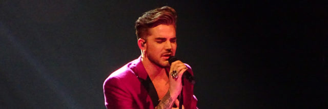 Adam Lambert performs onstage while wearing a purple suit.